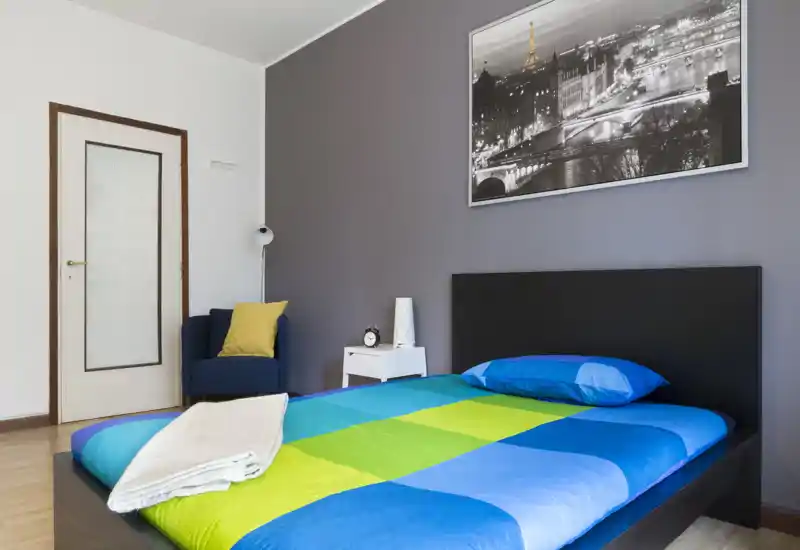 Bright, modern bedroom with a double bed dressed in vibrant blue and green bedding, a simple black headboard, bedside table with lamp and alarm clock, and a cozy armchair in the corner.