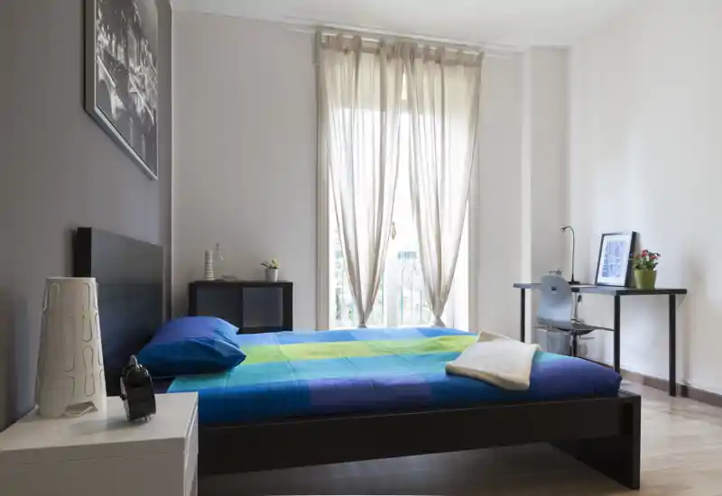 Bright, modern bedroom with a double bed, colorful bedding, natural light from tall window and a small workspace by the wall.