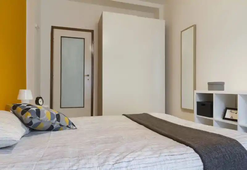 Bright, modern bedroom with a large bed dressed in neutral linens, a decorative pillow and a dark throw. Clean lines, warm accent wall and simple storage make the space feel welcoming and tidy — ideal for rental photos.