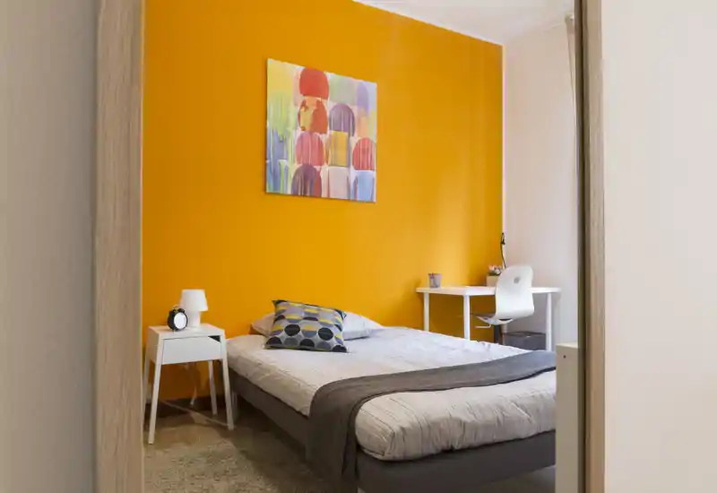 Sunny, modern bedroom with a bold orange accent wall, double bed dressed with neutral linens and a decorative pillow, and a small desk by the window — ideal for a bright, cozy stay.