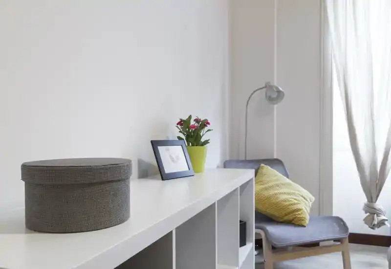 Bright, minimal living area with a comfortable armchair, floor lamp, decorative cushion and small plant on a modern shelving unit. Soft natural light and neutral tones create a calm, inviting corner ideal for reading or relaxing.