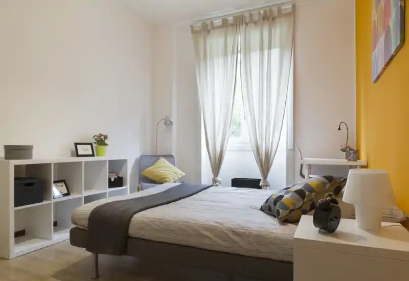 Dormitorio luminoso y acogedor con cama doble, textiles suaves y una gran ventana que aporta luz natural. Pared de acento cálida, mesita con lámpara y cojines decorativos que crean un ambiente agradable.