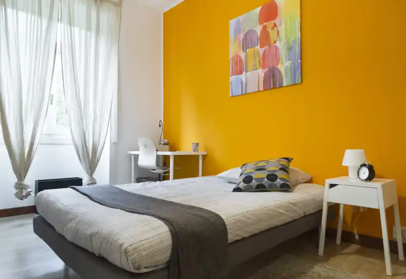 Dormitorio moderno y luminoso con cama doble cómoda, pared de acento colorida y un pequeño espacio de trabajo junto a la ventana. La luz natural y los textiles crean un ambiente acogedor perfecto para alquileres.