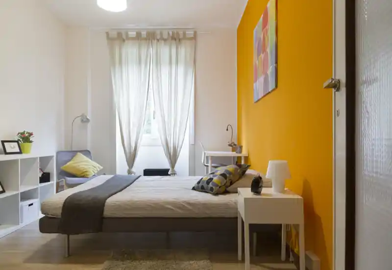 Dormitorio luminoso y bien decorado con cama doble junto a una pared naranja llamativa, luz natural suave desde la ventana, cojines decorativos y un pequeño espacio de trabajo al fondo.