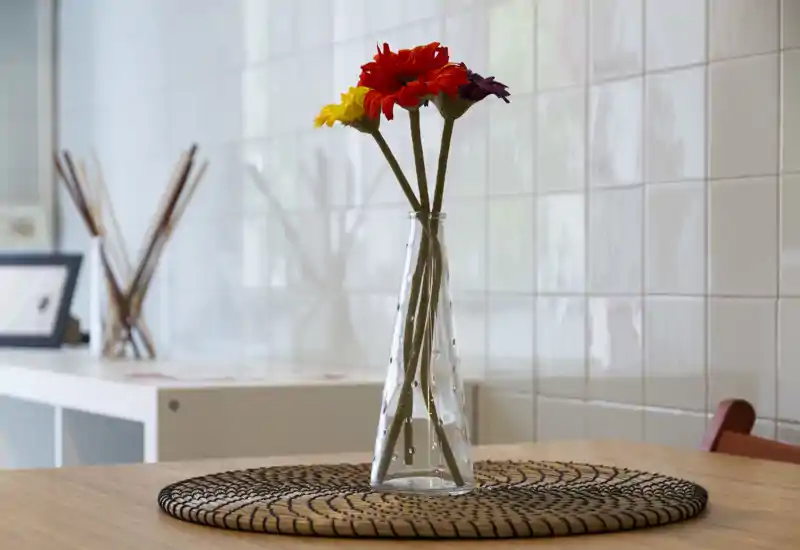 A close-up of a dining surface with a clear glass vase holding colorful flowers, placed on a woven placemat against a tiled wall — a simple, well-lit dining corner.