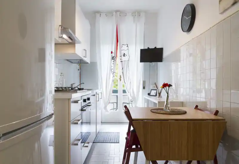 Bright, modern galley kitchen with white cabinets, a wooden dining table for two and natural light from a French door leading to a small balcony.