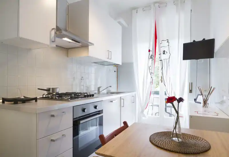 Bright, modern kitchenette with white cabinets, gas stove, built-in oven and a small dining table by the window with decorative flowers.