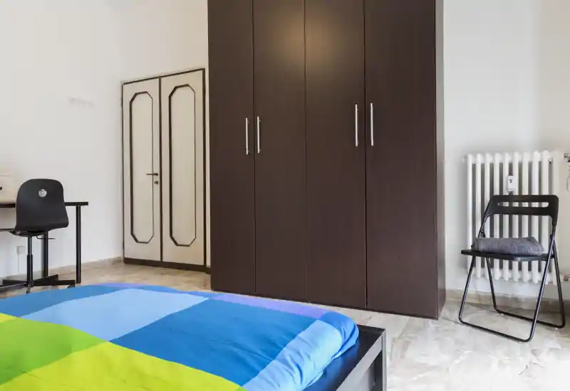 Bright, simple bedroom with a colorful bed cover, a large dark wardrobe, desk and chair — functional and tidy for a rental listing.