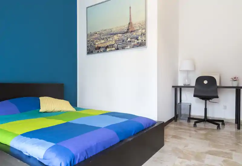 Bright, modern bedroom with a full bed dressed in colorful bedding and a small desk area — clean lines and pleasant natural light make this room inviting.