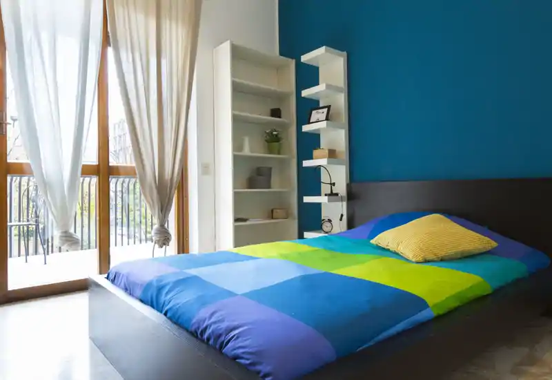 Bright, modern bedroom with a double bed dressed in colorful bedding, a tall shelving unit and large balcony doors providing abundant natural light.