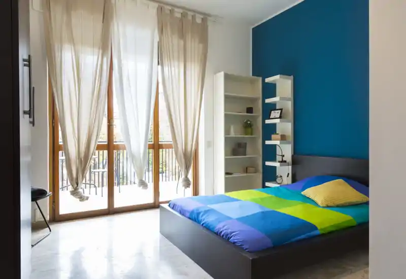 Bright, modern bedroom with a low platform bed dressed in colorful geometric bedding, a blue accent wall and large floor-to-ceiling windows letting in abundant natural light.