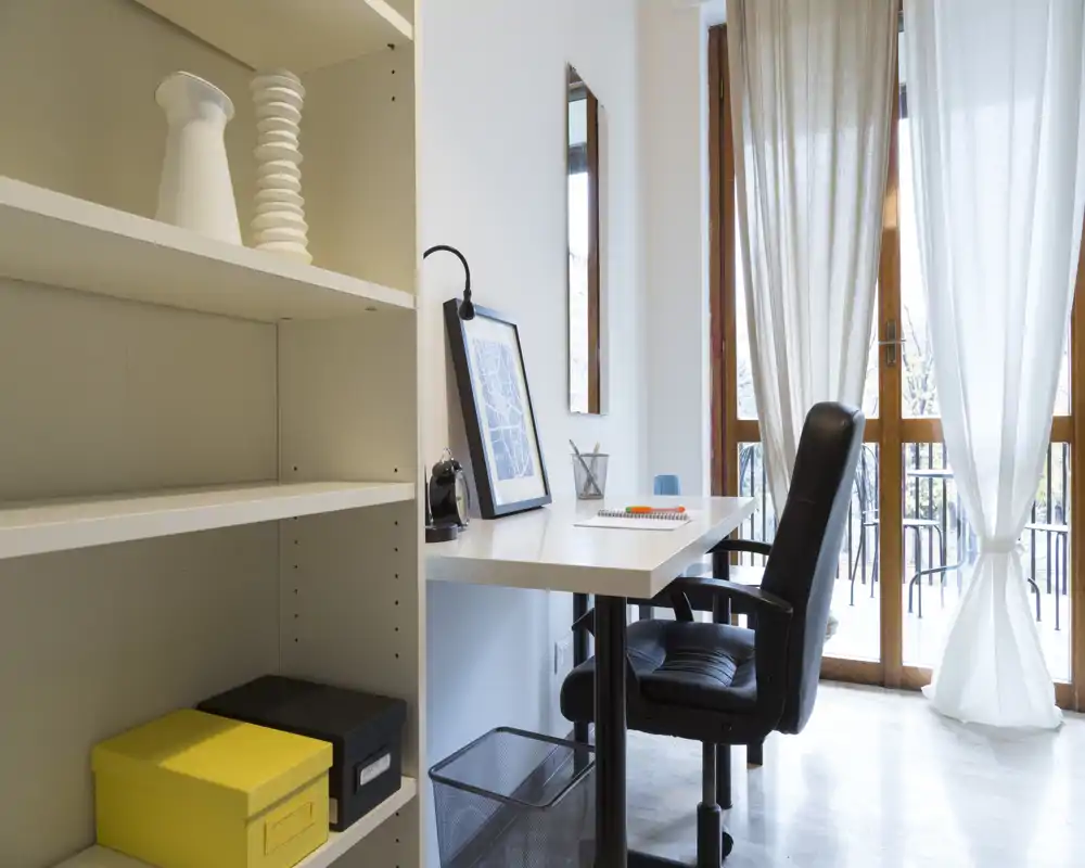 Angolo studio luminoso e minimal con scrivania bianca, sedia ergonomica nera e scaffalatura; luce naturale attraverso tende leggere verso un piccolo balcone.