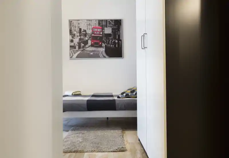 Camera da letto moderna e compatta con letto singolo ben rifatto, cuscino decorativo e piccolo tappeto; pareti neutre con quadro urbano per un aspetto minimal pulito.