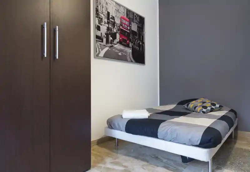 Piccola camera da letto moderna con letto singolo basso, cuscino decorato e armadio in legno scuro. Toni neutri e arredo minimal creano un ambiente luminoso e tranquillo, ideale per soggiorni brevi.