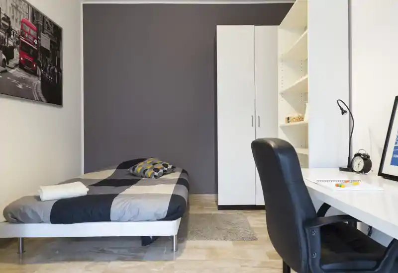 Camera singola moderna e compatta con letto ben rifatto, armadio e postazione di lavoro. Toni neutri e arredo essenziale creano uno spazio funzionale e tranquillo ideale per uno studente o un ospite singolo.