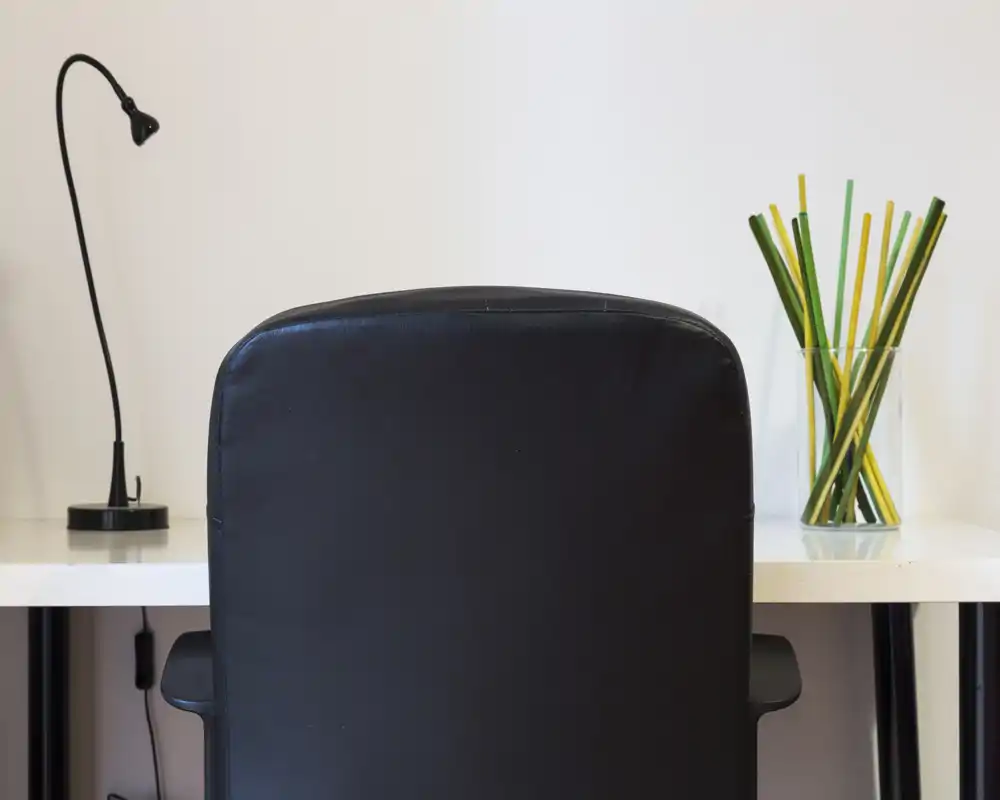 Simple workspace shot showing the back of a black office chair in front of a clean white desk with a small lamp and decorative glass vase with colored sticks.