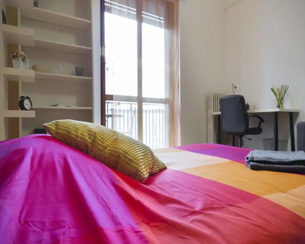 Bright, colorful bedroom with a large bed in the foreground, cozy cushion and natural light from a balcony window. A small desk and shelving provide functional space for work and storage.