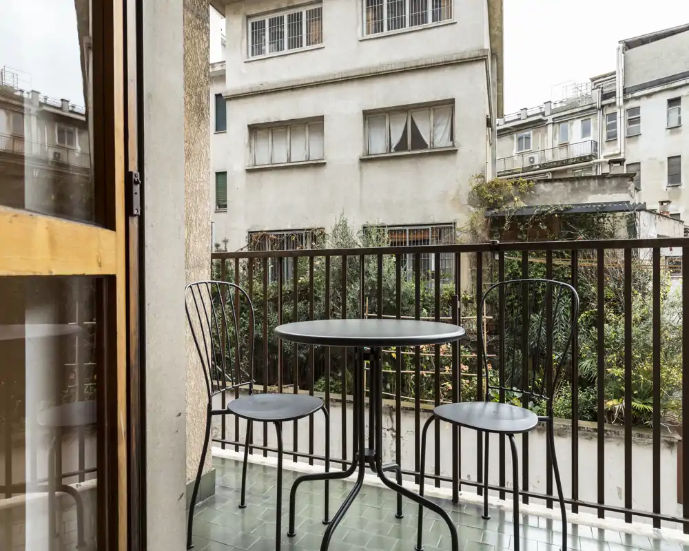 Sunny-feel balcony with a small round bistro table and two metal chairs overlooking a courtyard garden — ideal for morning coffee or a quiet outdoor break.