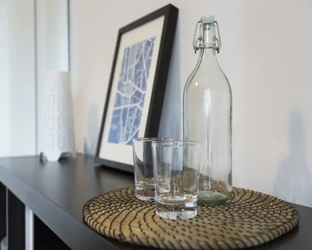 Close-up of a living-area surface with a decorative woven placemat, two clear glasses, and a tall glass bottle, set against framed artwork—clean, minimalist styling.