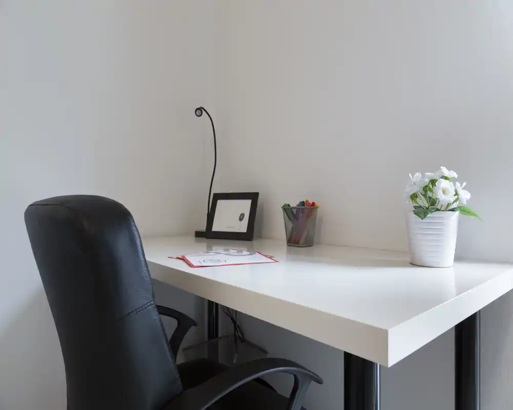 Bright, minimal desk corner with a white tabletop, black office chair, small lamp, framed photo, pen holder and a potted plant — a tidy living/working nook.