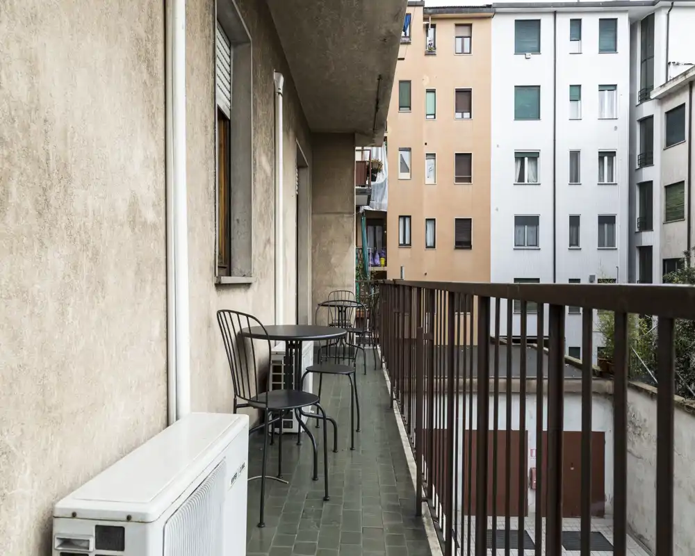 Long narrow balcony with metal railing, small round tables and chairs, and an outdoor air-conditioning unit; urban view of neighboring apartment façades.