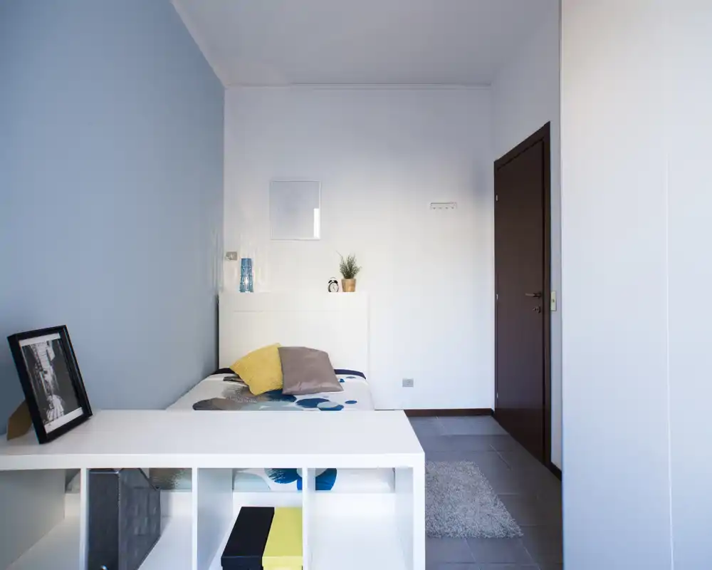 Camera da letto luminosa e compatta con un letto singolo, cuscini decorativi e un mobile contenitore moderno ai piedi del letto. Linee pulite e una parete azzurra creano un’atmosfera calma e ariosa.