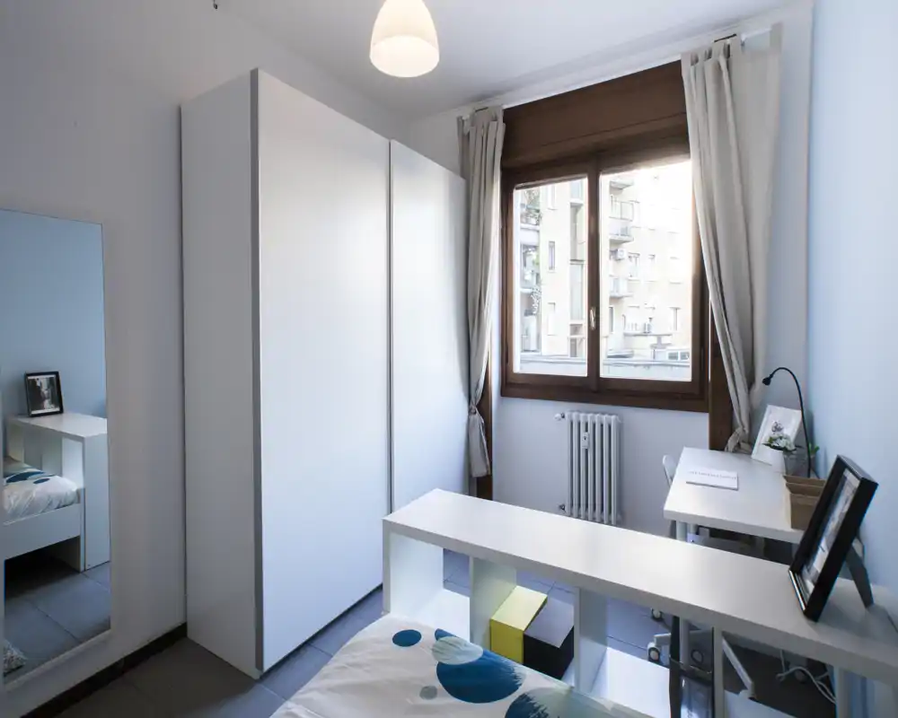 Camera singola moderna e luminosa con armadio bianco, scrivania vicino alla finestra e letto ordinato. La luce naturale illumina l’ambiente e l’arredo essenziale crea uno spazio contemporaneo e rilassante.