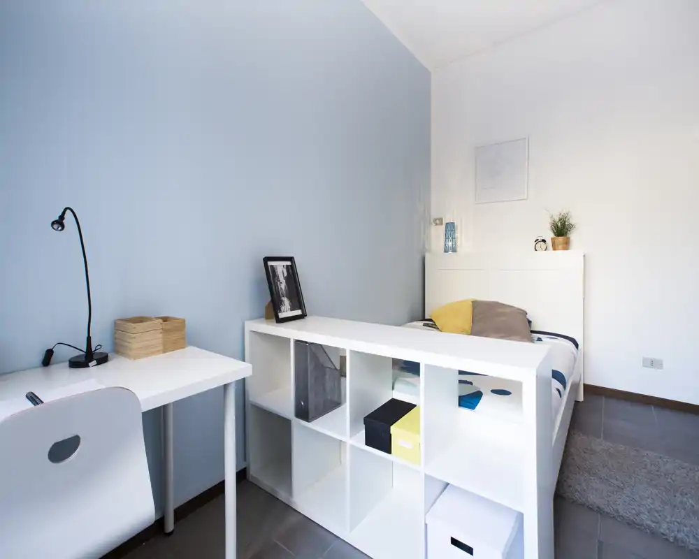 Camera singola moderna e luminosa con testiera e libreria bianca alla fine del letto, scrivania semplice e arredamento minimal — ideale per studenti o affitti brevi.