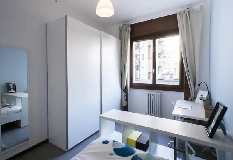 Camera singola moderna e luminosa con armadio bianco, scrivania vicino alla finestra e letto ordinato. La luce naturale illumina l’ambiente e l’arredo essenziale crea uno spazio contemporaneo e rilassante.