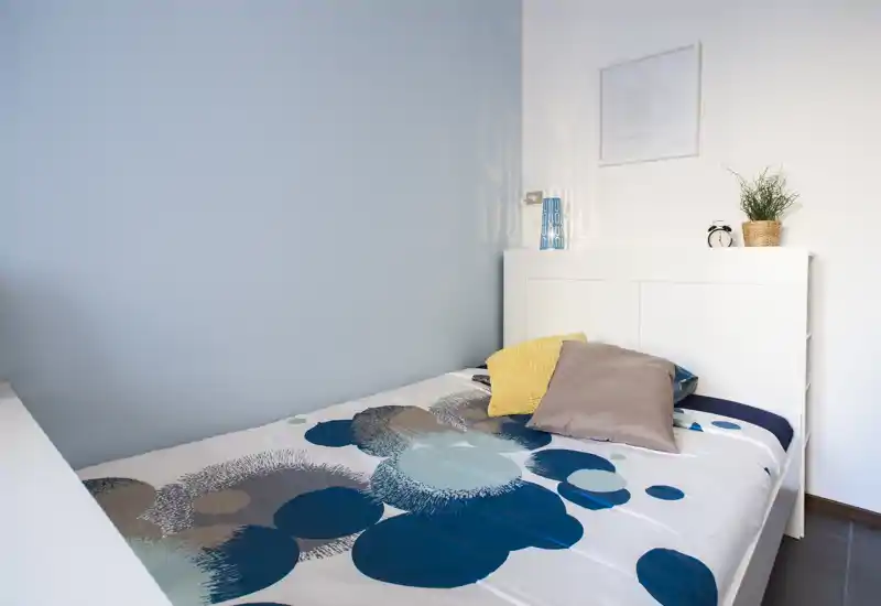 Camera da letto ordinata e luminosa con letto completo, cuscini decorativi e una testiera semplice; parete accento azzurra e arredamento minimale creano un’atmosfera calma.