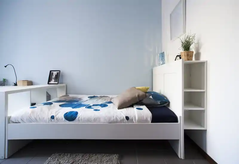 Camera da letto luminosa e minimal con struttura letto bianca, copriletto a motivi e una testiera con ripiani; arredo moderno e luce naturale soffusa.
