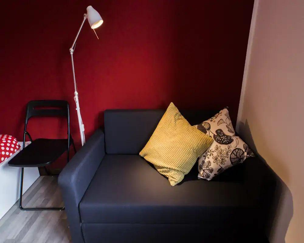 Piccolo soggiorno con divano scuro, cuscini colorati e lampada da terra davanti a una parete rossa: accogliente e ben arredato per un appartamento compatto.