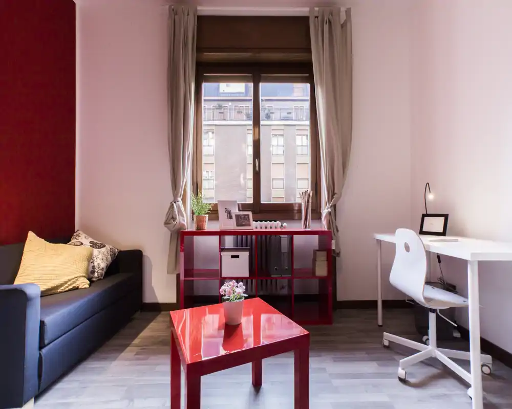 Zona giorno moderna e luminosa con divano confortevole, cuscini decorativi, un tavolino rosso e una finestra che garantisce luce naturale — include un piccolo angolo studio.