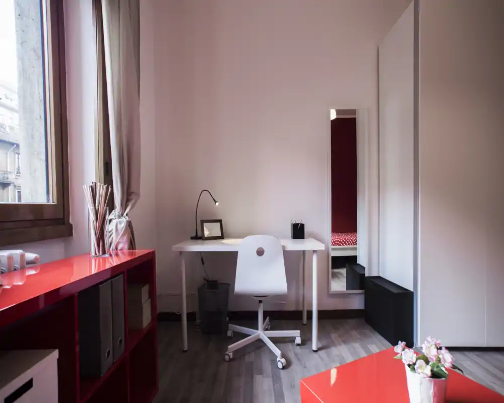 Camera da letto compatta con una scrivania minimalista e uno specchio alto che riflette un letto con copertura rossa a pois; luce naturale da una finestra che illumina le pareti rosa e il pavimento in legno.