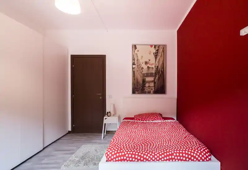 Camera singola compatta e di design con letto bianco e copripiumino rosso a pois, comodino e quadro alle pareti; linee pulite e parete rossa a contrasto creano un ambiente moderno e accogliente.