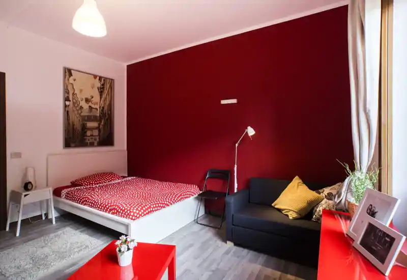 Camera da letto luminosa e di stile con letto bianco completo e parete rossa come elemento d'accento. Presenti un piccolo divano nero, comodini e luce naturale da una grande finestra.