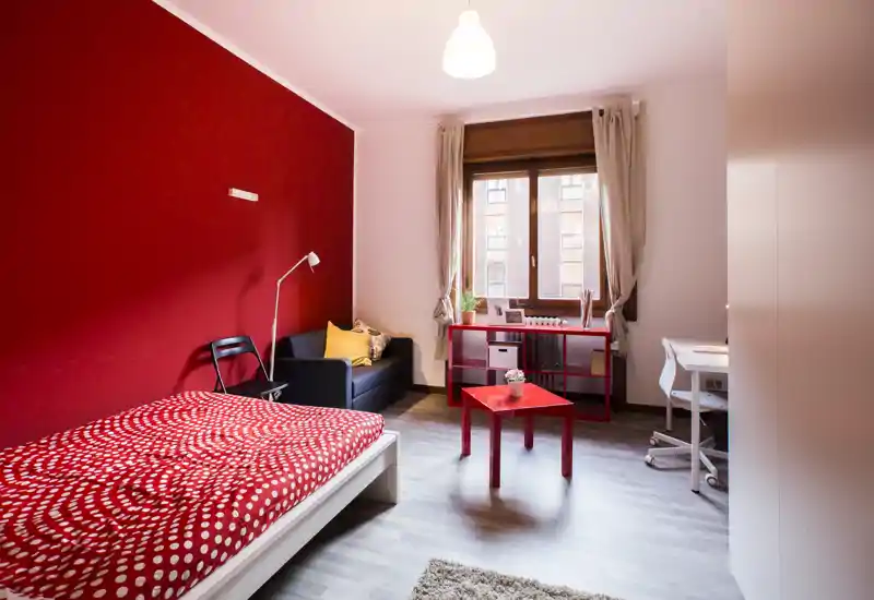Camera da letto luminosa e moderna con parete rossa, ampia finestra e letto singolo con copriletto rosso a pois; arredata con piccolo divano, scrivania e scaffale — ideale per studente o occupante singolo.