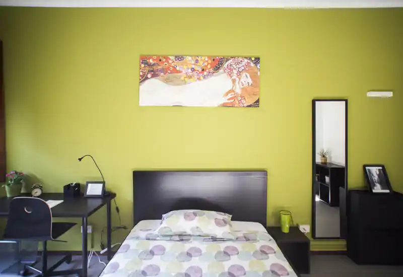Camera da letto luminosa e moderna con un letto singolo ben rifatto, quadro decorativo e una piccola scrivania a sinistra. La stanza presenta una parete verde a contrasto, uno specchio a figura intera e spazio contenitivo ordinato — perfetta per affitti brevi o studenti.