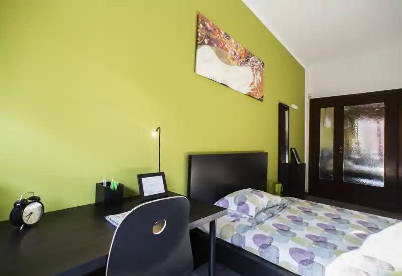 Camera da letto moderna e luminosa con letto singolo dal copriletto decorato, testiera nera e una scrivania su una parete verde. La stanza è ordinata e accogliente, con buona luce naturale e dettagli decorativi semplici.