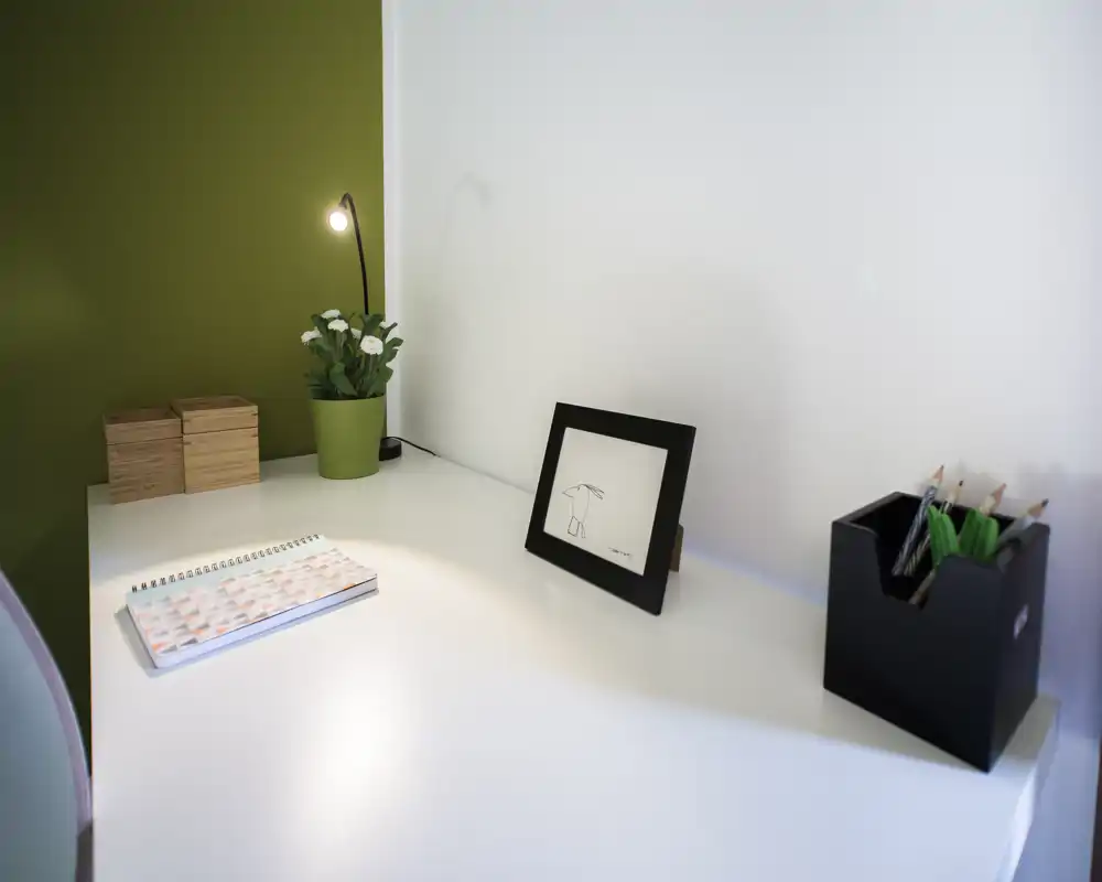 Angolo studio luminoso e minimal in una zona giorno con scrivania bianca pulita, lampada, cornice con disegno, pianta e portapenne — ideale per lavoro concentrato.
