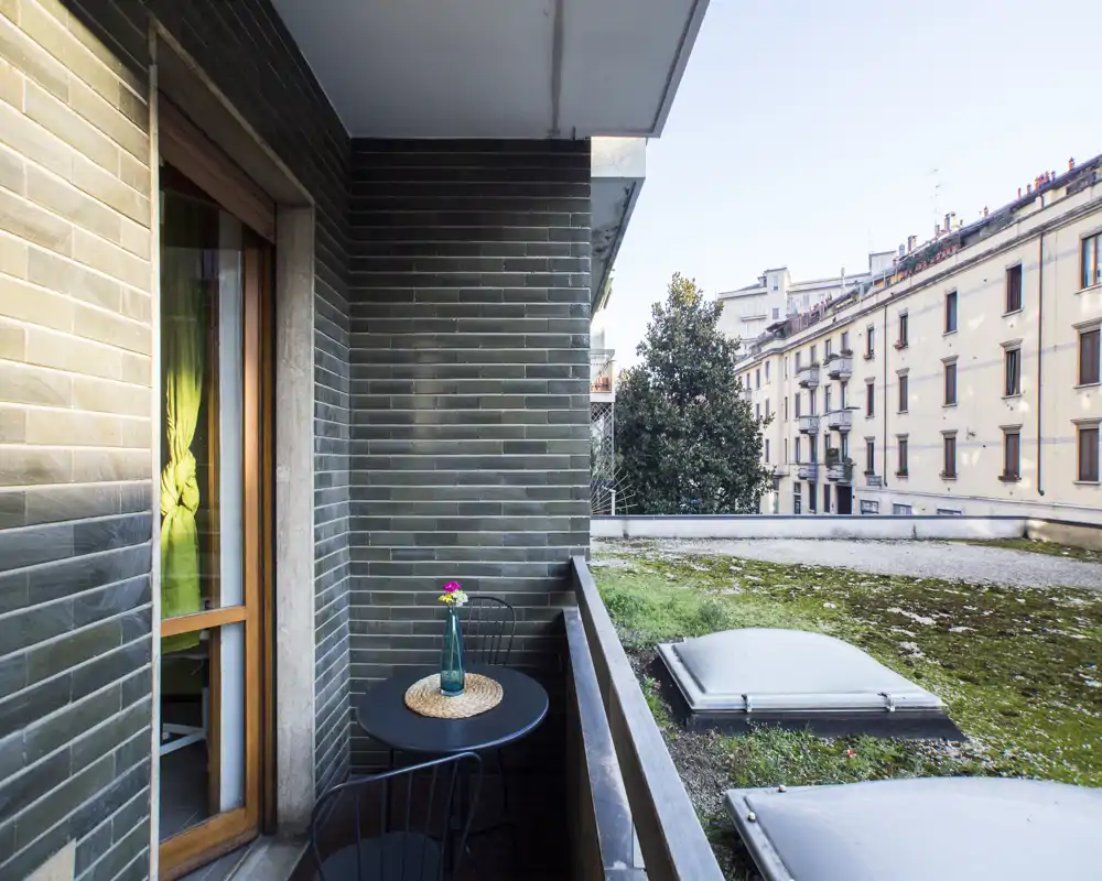 Piccolo balcone urbano con un tavolino rotondo compatto, due sedie in metallo e un fiore in vaso; vista su un tetto piano e gli edifici residenziali vicini.