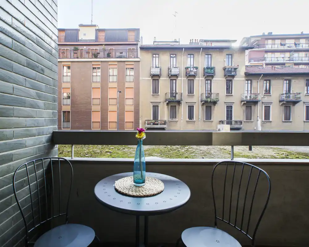 Accogliente balcone con piccolo tavolo rotondo, due sedie in metallo e una bottiglia decorativa — perfetto per un caffè mattutino con vista sugli edifici.