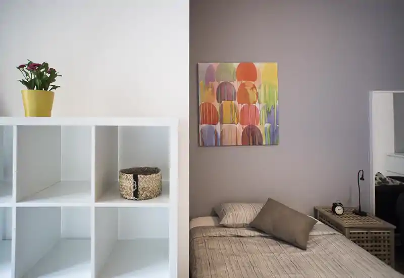 Angolo camera luminoso e curato con letto singolo, biancheria a righe e comodino con lampada e sveglia. Un quadro astratto colorato e una libreria bianca con cesta decorativa e pianta aggiungono personalità.
