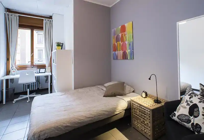 Camera da letto luminosa e ordinata con letto matrimoniale, comodino e area studio vicino alla finestra; biancheria neutra e un quadro colorato rendono l'ambiente accogliente.
