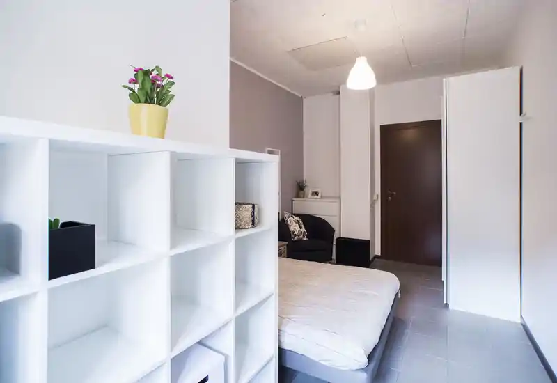 Camera da letto moderna e luminosa con letto ben fatto, divisorio a scaffali bianco, una poltrona accogliente e luce soffusa dall'alto — arredo essenziale e ordinato, perfetto per annunci.