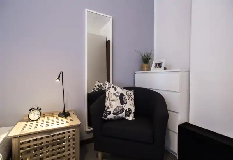 Angolo camera accogliente con poltrona, cuscino decorativo e comodino; pareti in toni neutri e uno specchio a figura intera creano un’atmosfera tranquilla.