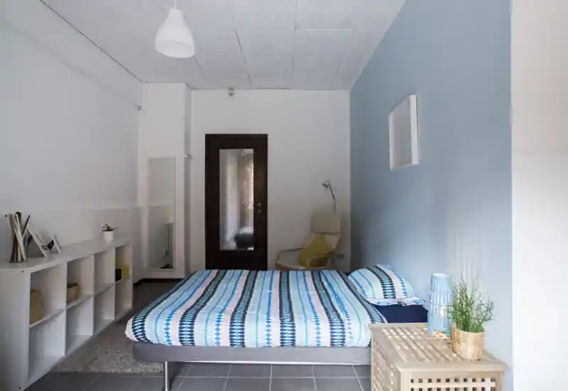 Camera da letto luminosa e ordinata con letto matrimoniale con copriletto a righe blu, comodino con pianta, una poltrona per la lettura vicino al muro e uno scaffale ordinato — moderna e tranquilla.