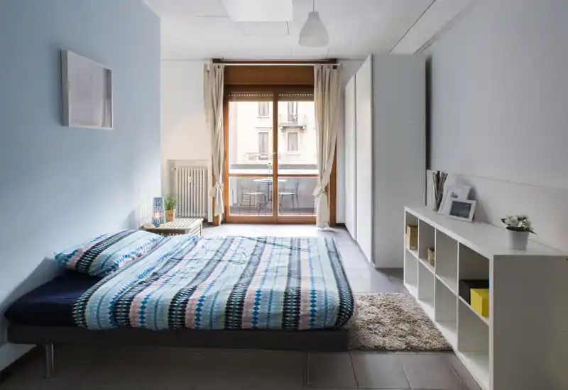Camera da letto moderna e luminosa con letto matrimoniale, biancheria a fantasia, tappeto, mobile contenitore e balcone che illumina naturalmente la stanza. Linee pulite e colori morbidi rendono l’ambiente accogliente e ideale per affitti.