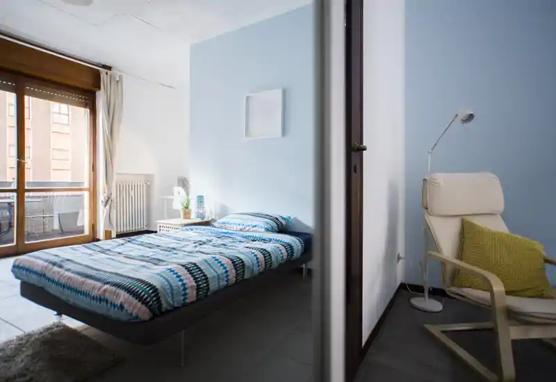 Camera da letto luminosa e ben arredata con letto matrimoniale, comodino e una poltrona vicino alla finestra. La grande porta finestra assicura molta luce naturale e le tonalità pastello creano un’atmosfera rilassante.