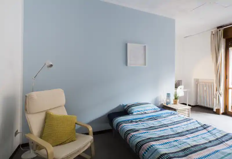 Camera singola luminosa e ordinata con letto basso con biancheria blu a motivo, poltrona comoda con cuscino giallo, comodino e luce naturale dalla porta finestra.